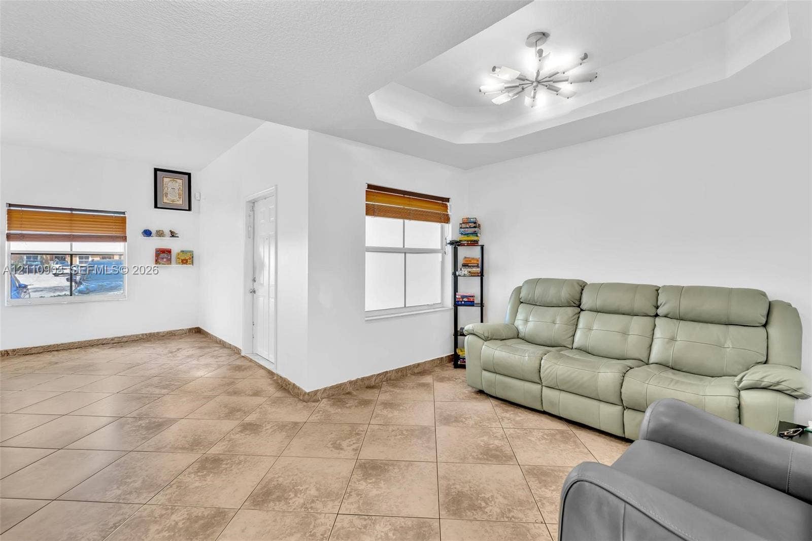 Interior space featuring large-format tile flooring, a recessed tray ceiling, a multi-arm modern chandelier, and two windows with integrated blinds