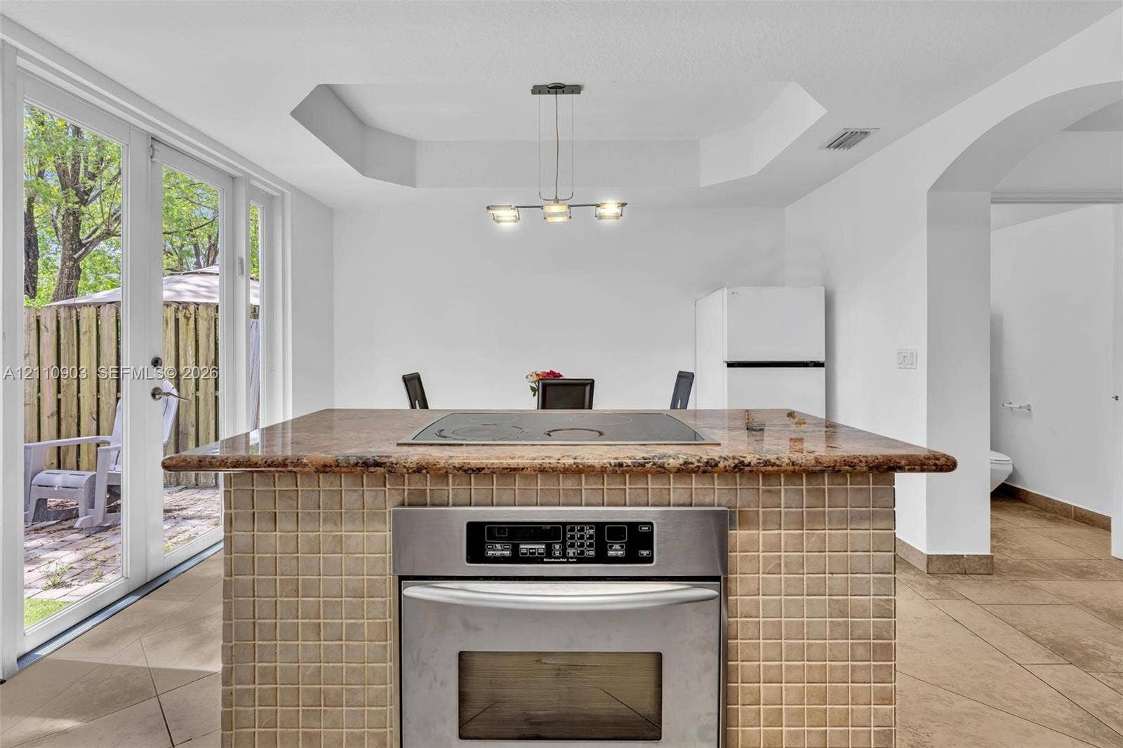 Kitchen island featuring a cooktop, built-in stainless steel oven, stone countertop, and tile-finish base, complete with a coffered ceiling and glass paneled doors