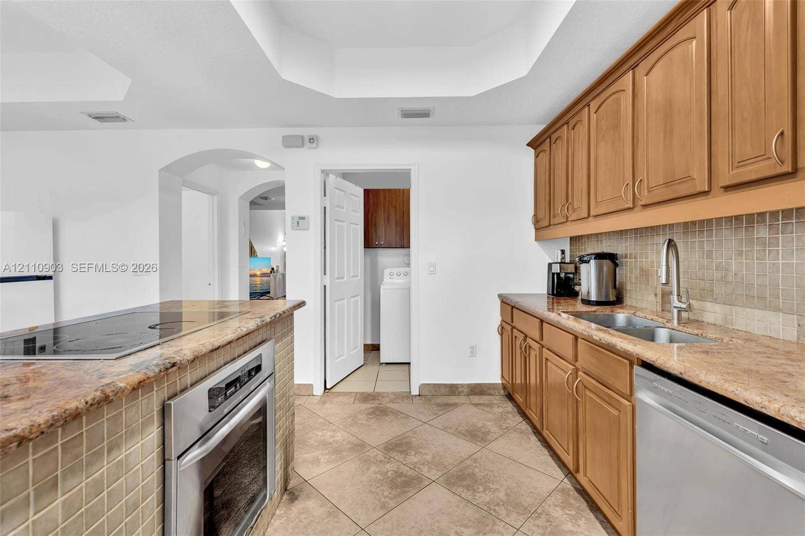 Kitchen featuring honey-toned wood cabinetry, stone countertops, stainless steel wall oven, mosaic tile backsplash, and tray ceiling