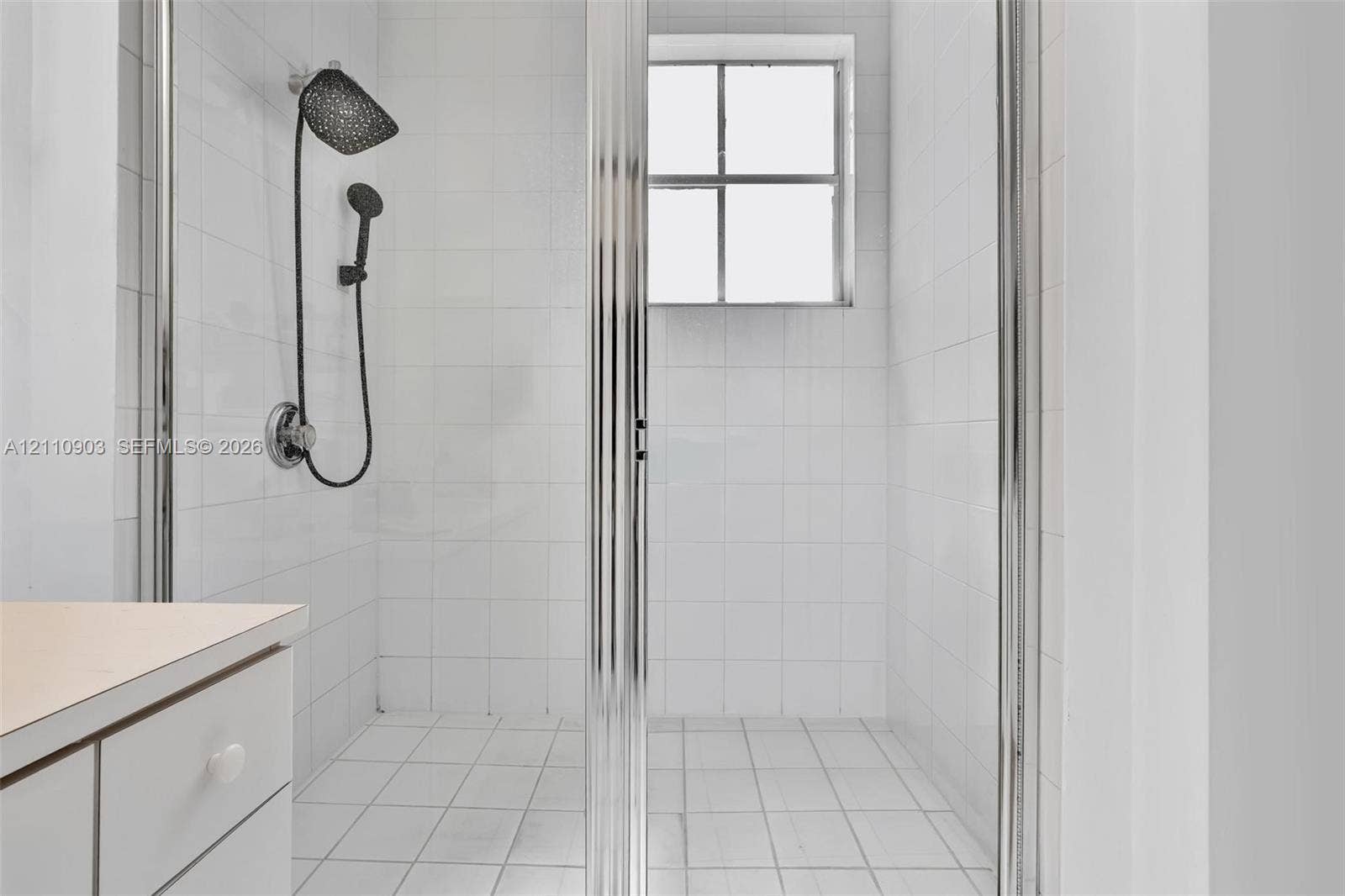 Walk-in shower enclosure with white tiled walls, square-grid tile flooring, chrome-framed glass door, and a dual-head shower fixture