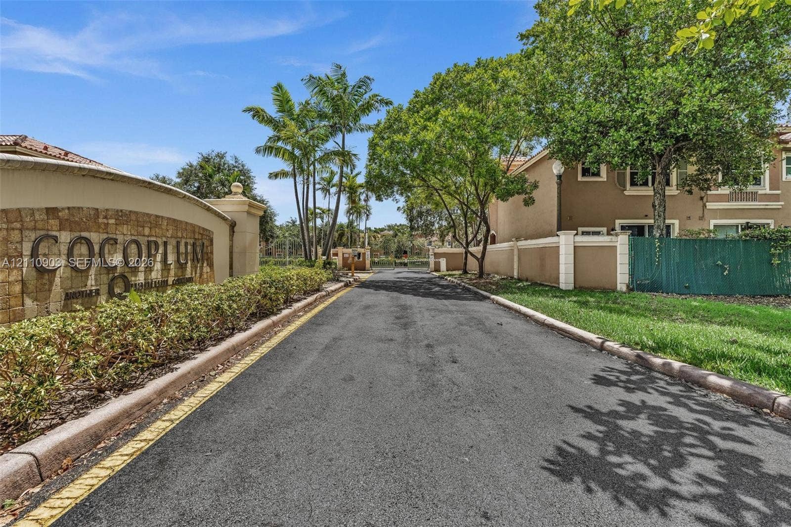 Gated entrance featuring a stone-clad community monument sign, paved driveway, perimeter wall with pilasters, and mature landscaping