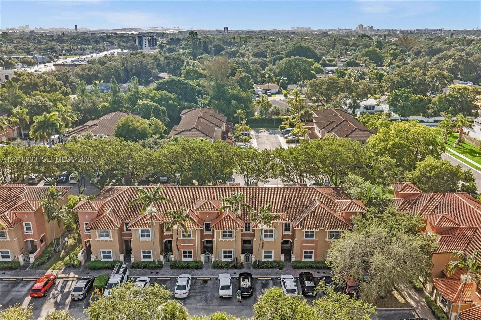 Aerial perspective of a multi-unit residential complex featuring a terracotta-style tile roof, stucco exterior, assigned parking, and mature surrounding tree canopy