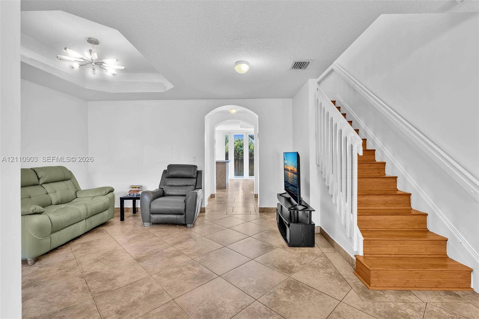 Tiled flooring, tiered tray ceiling with modern chandelier, staircase with white balusters and wood treads, and arched doorway