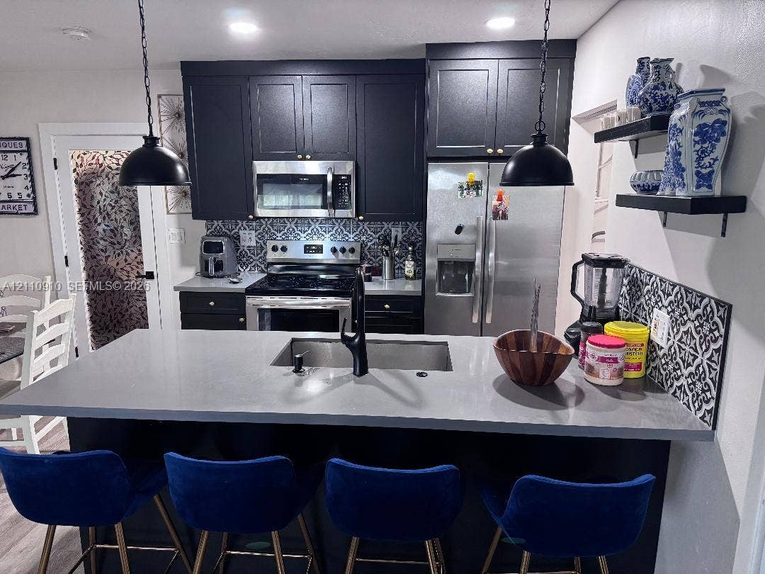 Kitchen featuring matte black cabinetry, solid surface countertop with an integrated stainless steel sink, patterned tile backsplash, stainless steel appliances, and dual pendant lighting