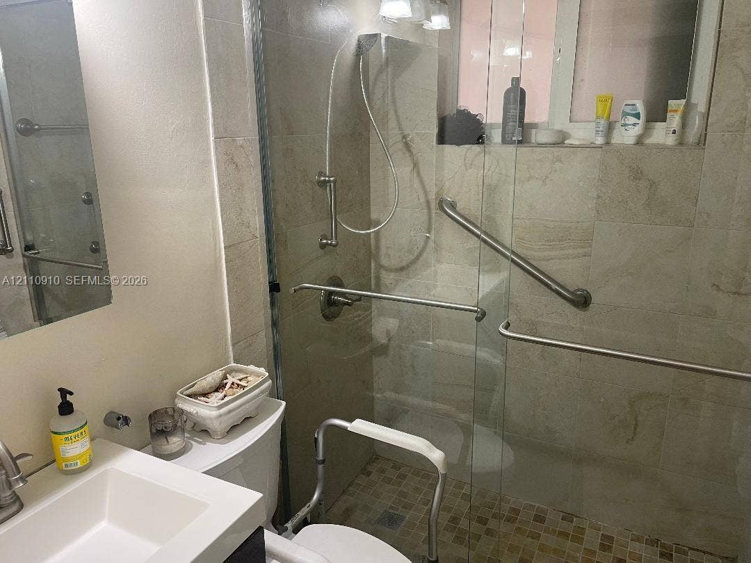 Walk-in shower featuring large-format tile surround, glass enclosure, and metallic grab bars, paired with a white rectangular basin sink and neutral wall-mounted lighting