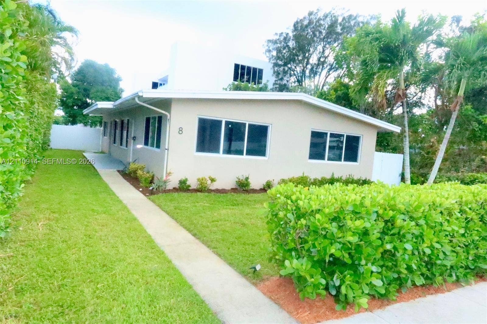 Stucco exterior residence featuring a concrete walkway, horizontal slider windows, and perimeter privacy hedges