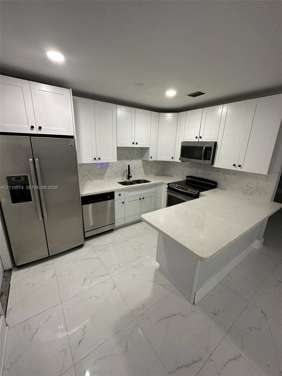 Kitchen featuring white shaker-style cabinetry, stone-finish countertops, stainless steel appliances, a peninsula, and large-format tile flooring with veining