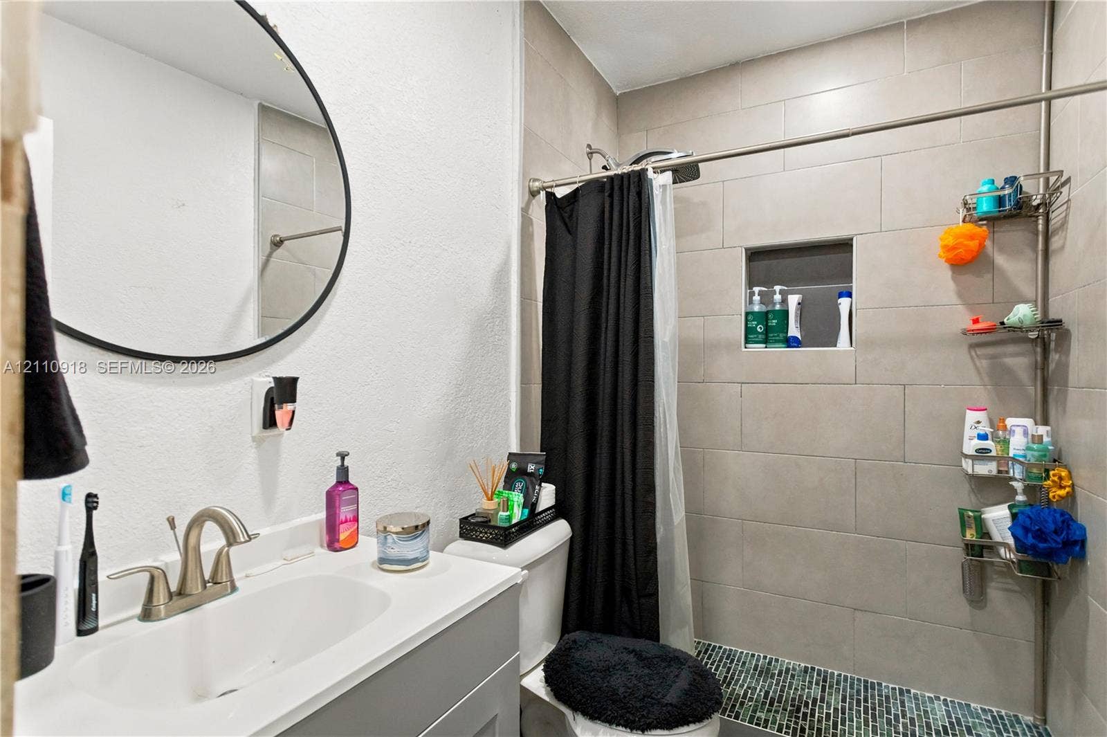 2007 NW 89th Street, Miami, FL 33147 Bathroom featuring a vanity with a white countertop and integrated sink, matte-finish faucet, circular wall mirror, and a shower area with tiled walls and a recessed niche