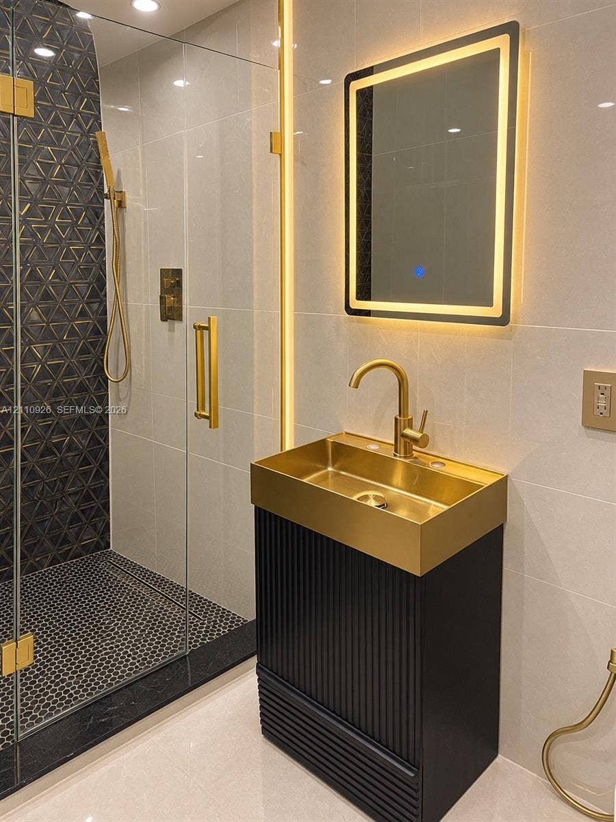 Fluted black vanity with integrated gold-tone sink, matching gold-tone hardware, LED-backlit mirror, and walk-in shower with geometric accent tiling