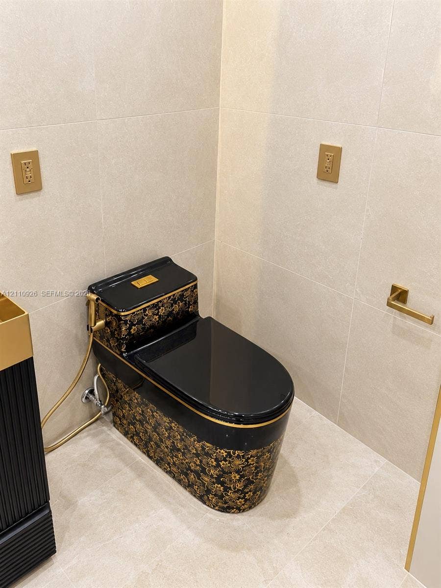 Custom black and gold patterned toilet featuring a matching bidet sprayer, complemented by light-toned large-format wall and floor tile, gold-finish electrical faceplates, and a wall-mounted gold-finish paper holder