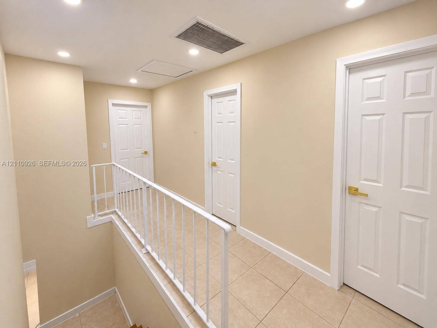 Hallway featuring tile flooring, six-panel interior doors with brass-tone hardware, recessed ceiling lighting, and a white metal railing