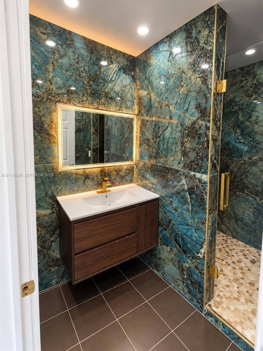 Floating vanity with a white countertop and gold-toned faucet, accented by full-height stone-finish wall cladding, recessed lighting, and a glass-enclosed shower with pebble-textured flooring