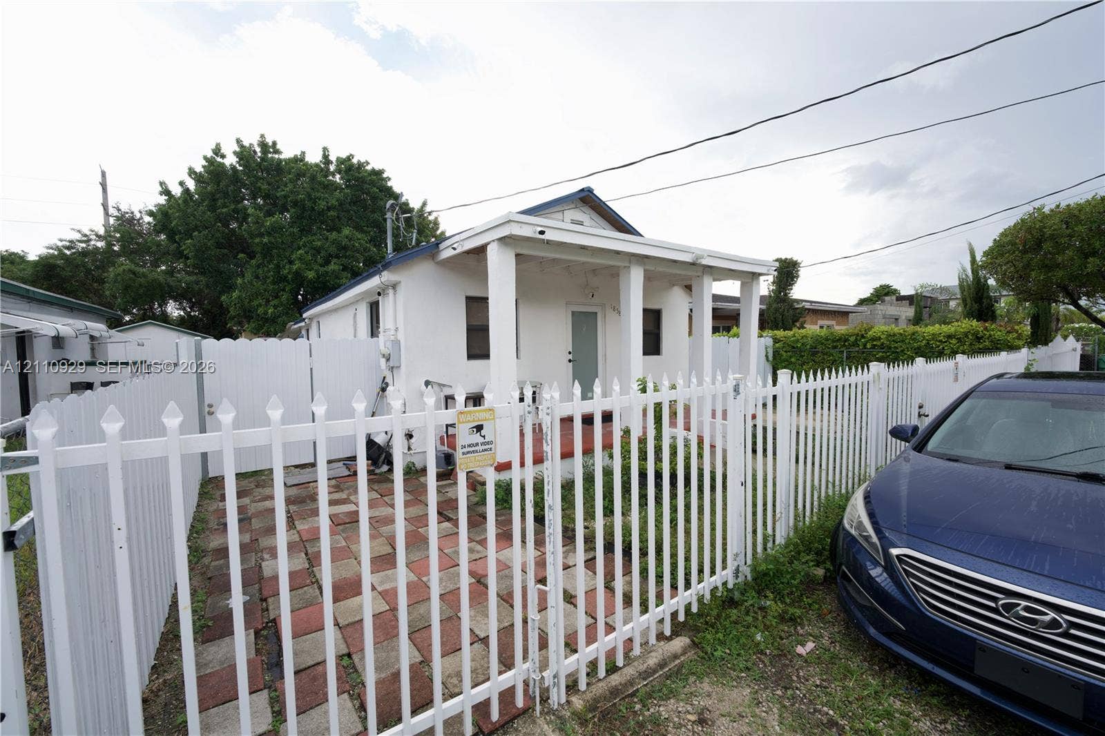Exterior featuring a covered front porch with square columns, a gated entry, a white vinyl perimeter fence, and a paved brick-style patio