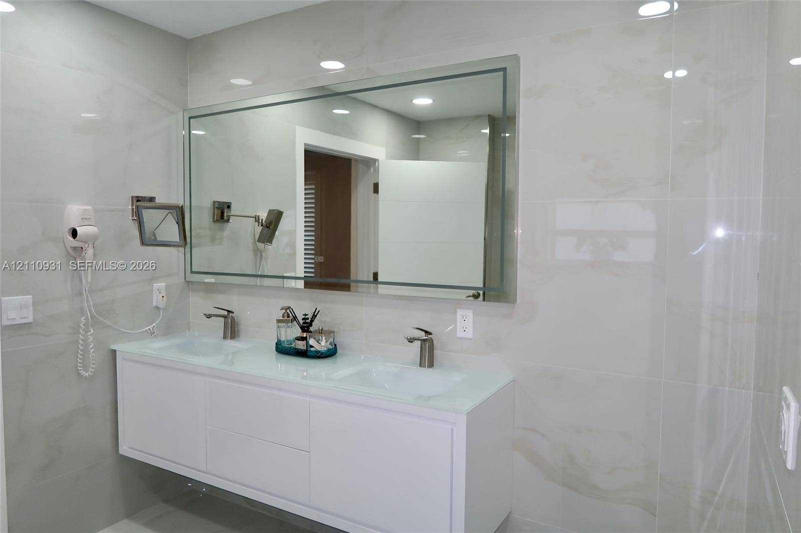 3005 SW 99th Court, Miami, FL 33165 Wall-mounted white dual-sink vanity featuring a glass countertop, large rectangular mirror with integrated lighting, wall-mounted adjustable magnification mirror, and large-format neutral-toned tile wall surfacing