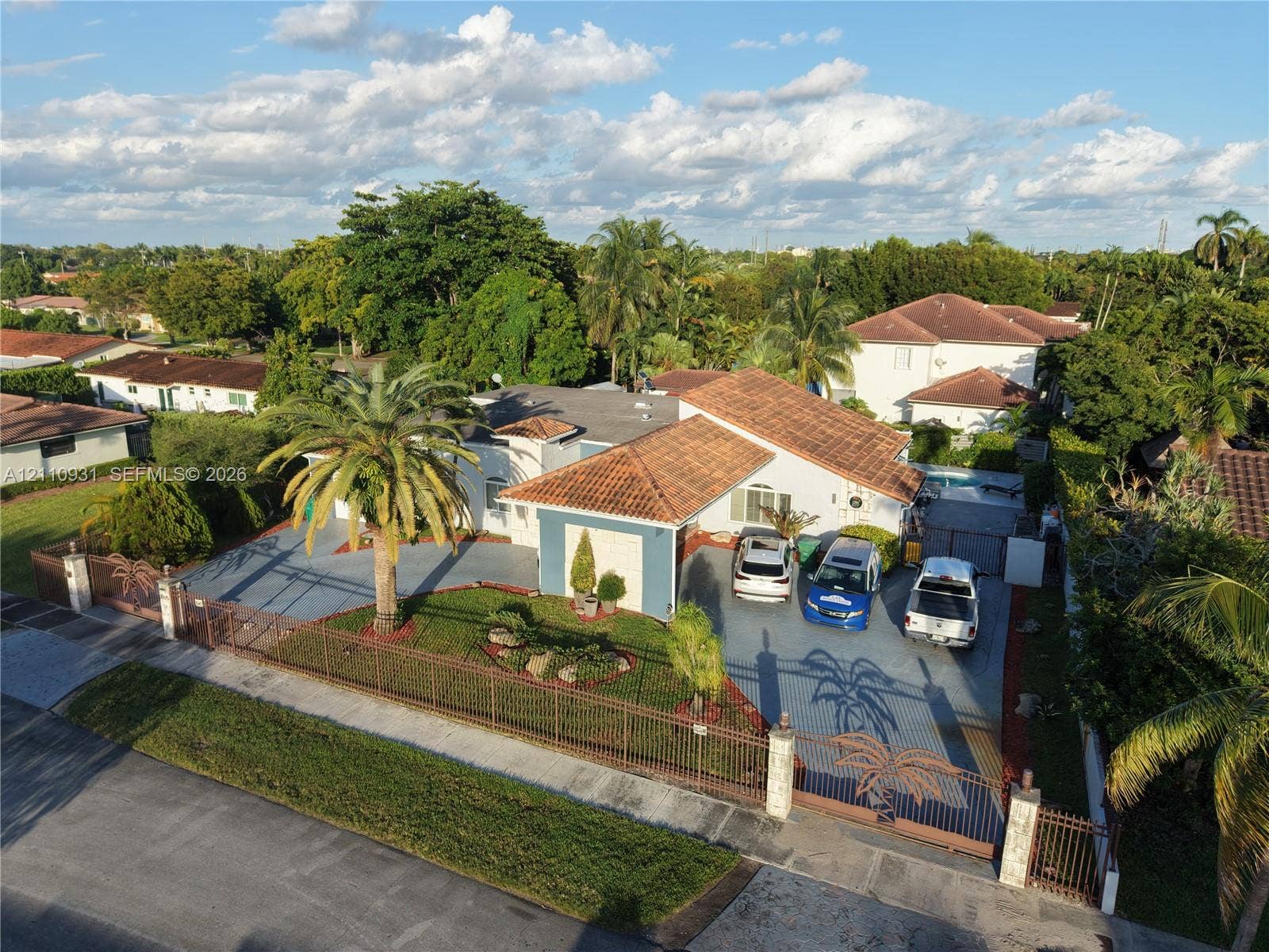 3005 SW 99th Court, Miami, FL 33165 Single-story residence featuring a clay tile roof, expansive paved driveway, decorative metal perimeter fencing, and mature landscaping