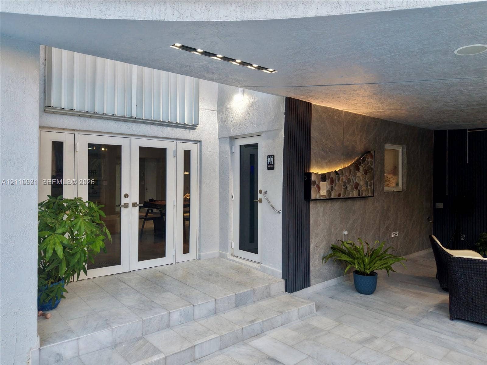 3005 SW 99th Court, Miami, FL 33165 Covered exterior entryway featuring stone-tiled flooring, double glass-panel doors, accent stone wall cladding with integrated lighting, and vertical slat detailing