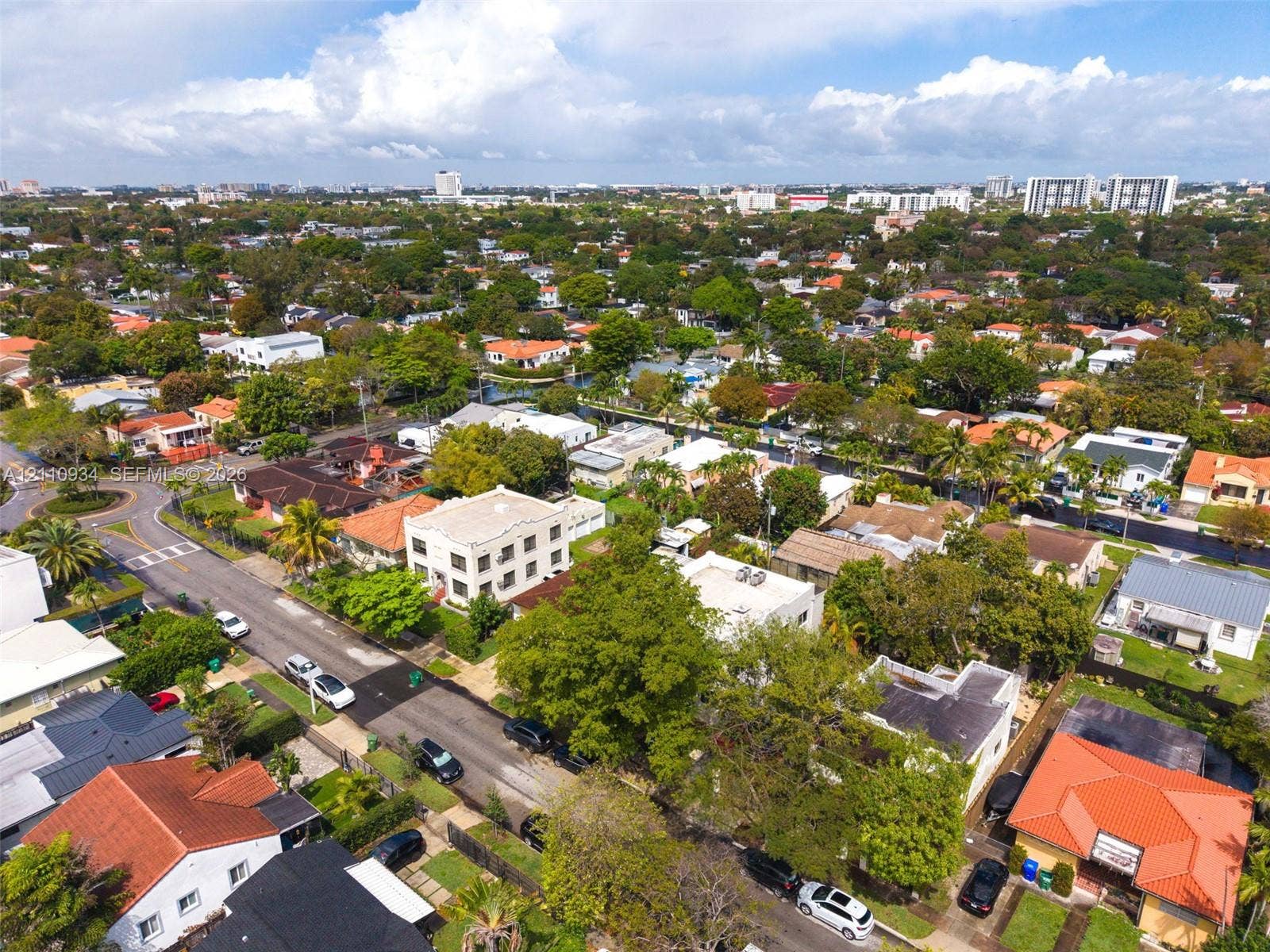 Aerial perspective of a multi-story residential structure featuring a flat roofline, stucco exterior, and multi-paned windows, situated within a dense urban landscape characterized by mature trees and established street grids