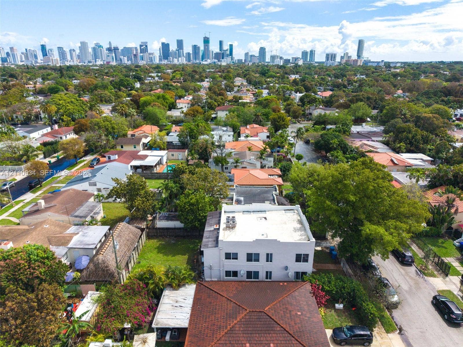 Aerial perspective of a multi-story building featuring a flat roof and surrounding mature landscaping, positioned within a suburban environment with a prominent city skyline backdrop