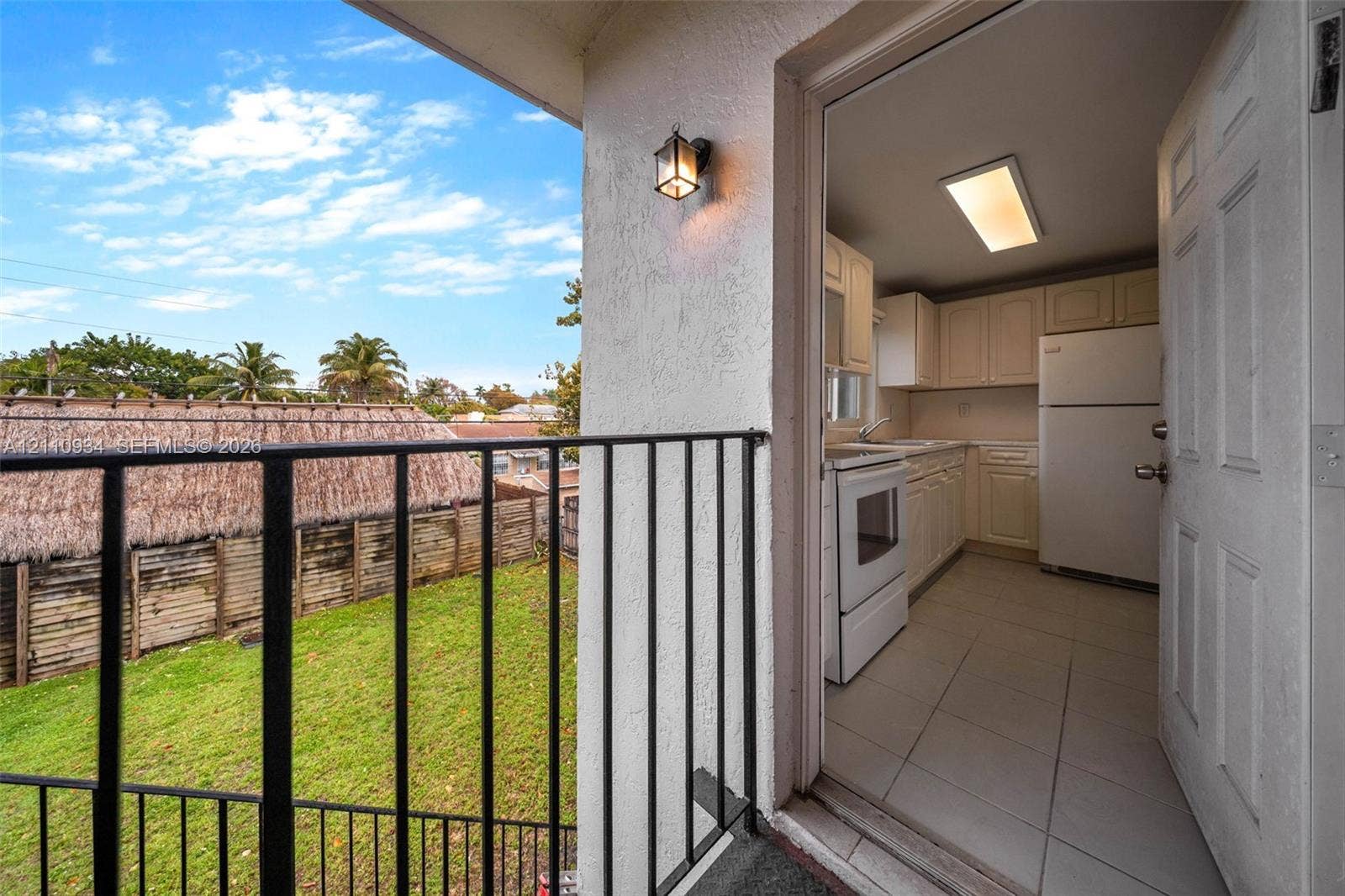 Balcony featuring a black metal railing and exterior light fixture, opening into a kitchen with light-toned cabinetry, tile flooring, and integrated white appliances