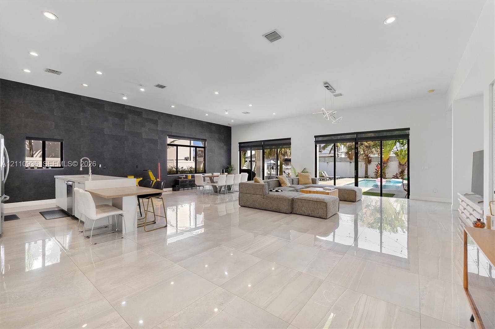 Open-concept living space featuring high-gloss tile flooring, recessed lighting, a statement dark tile accent wall, an island kitchen with built-in cabinetry, and multi-panel glass sliding doors