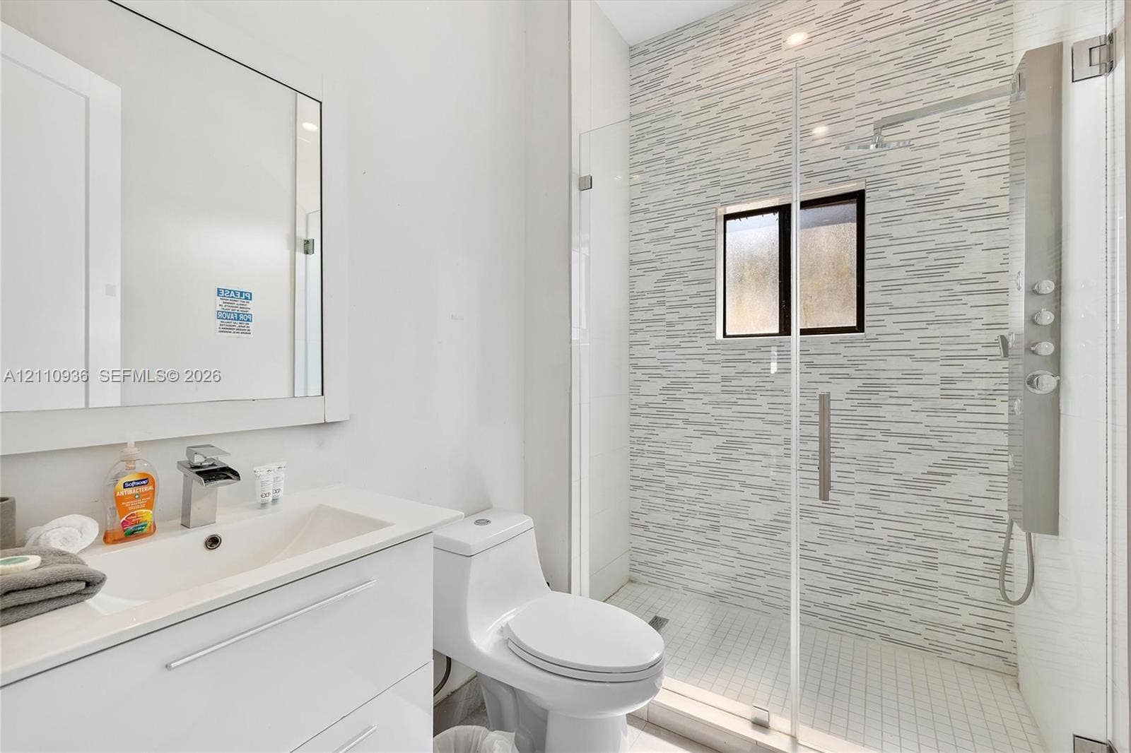 Modern bathroom featuring a white vanity with an integrated countertop, wall-mounted mirror, walk-in shower with a glass enclosure, patterned tile shower wall, and an integrated shower panel system