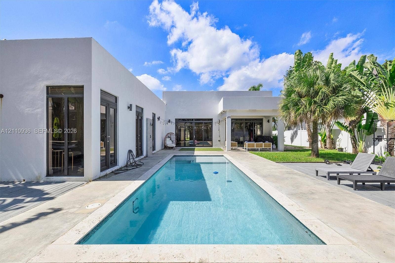 Modern exterior with smooth stucco finish, rectangular swimming pool with stone coping, concrete patio, covered outdoor area, and established palm trees