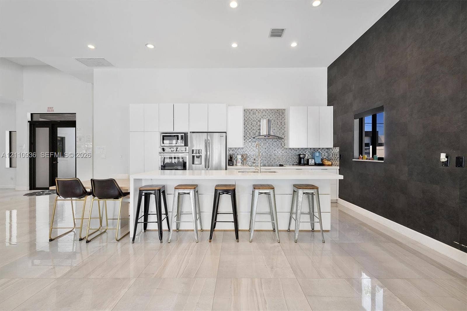 Modern kitchen featuring flat-panel white cabinetry, a waterfall-edge kitchen island, stone-finish tile flooring, a mosaic tile backsplash, and a dark tile accent wall