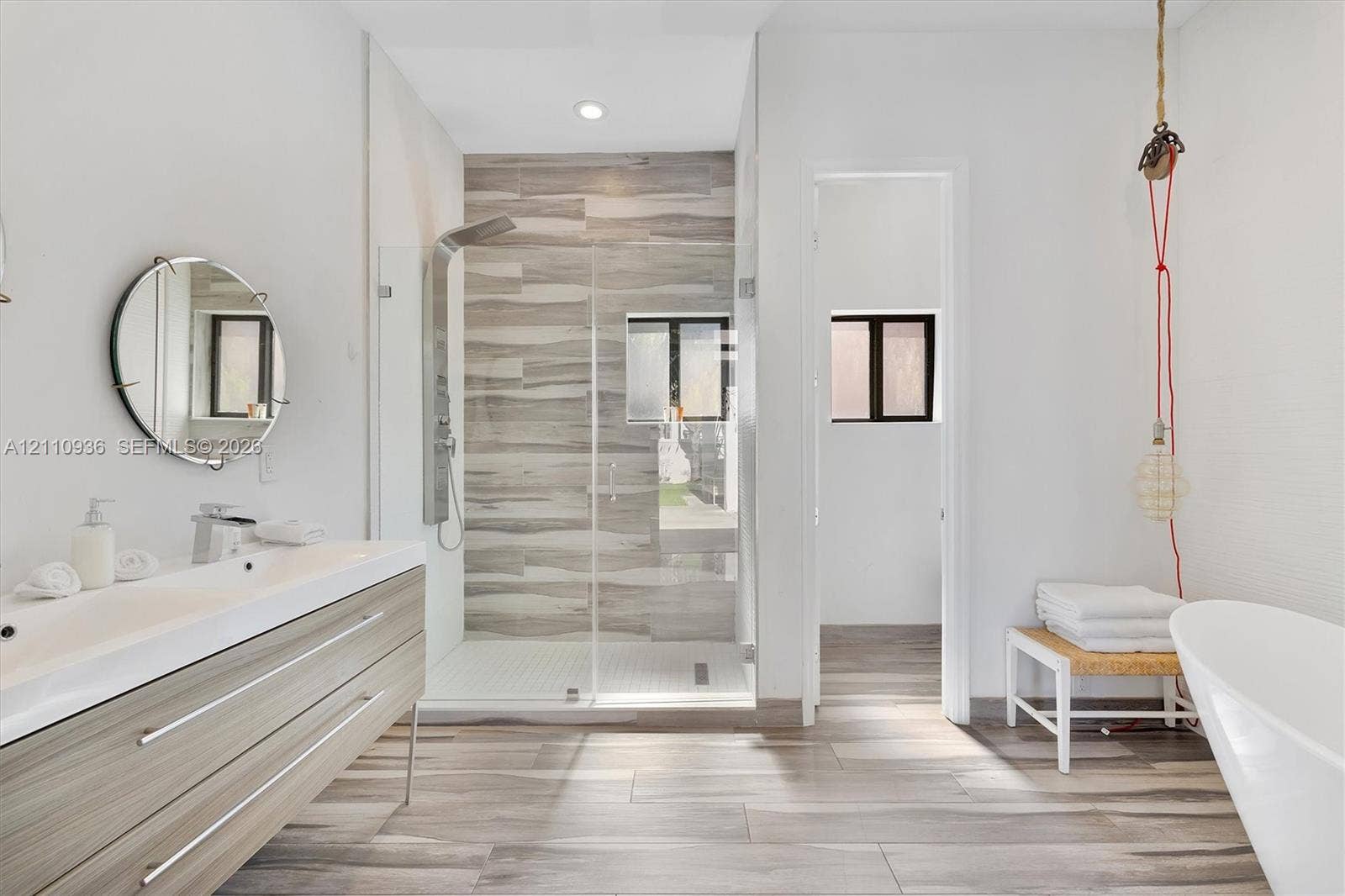 Modern floating vanity with a double-basin integrated countertop, walk-in glass-enclosed shower featuring horizontal tile accents, wall-mounted shower panel, wood-finish flooring, and a freestanding soaking tub