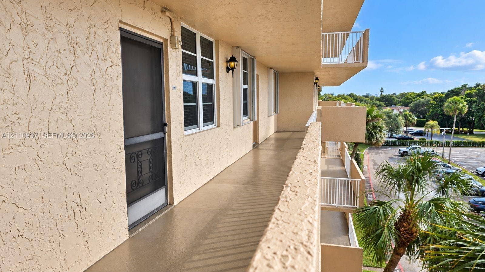 Exterior walkway featuring a stucco facade, metal screen door, white-framed multi-pane windows, wall-mounted coach lighting, and a white railing