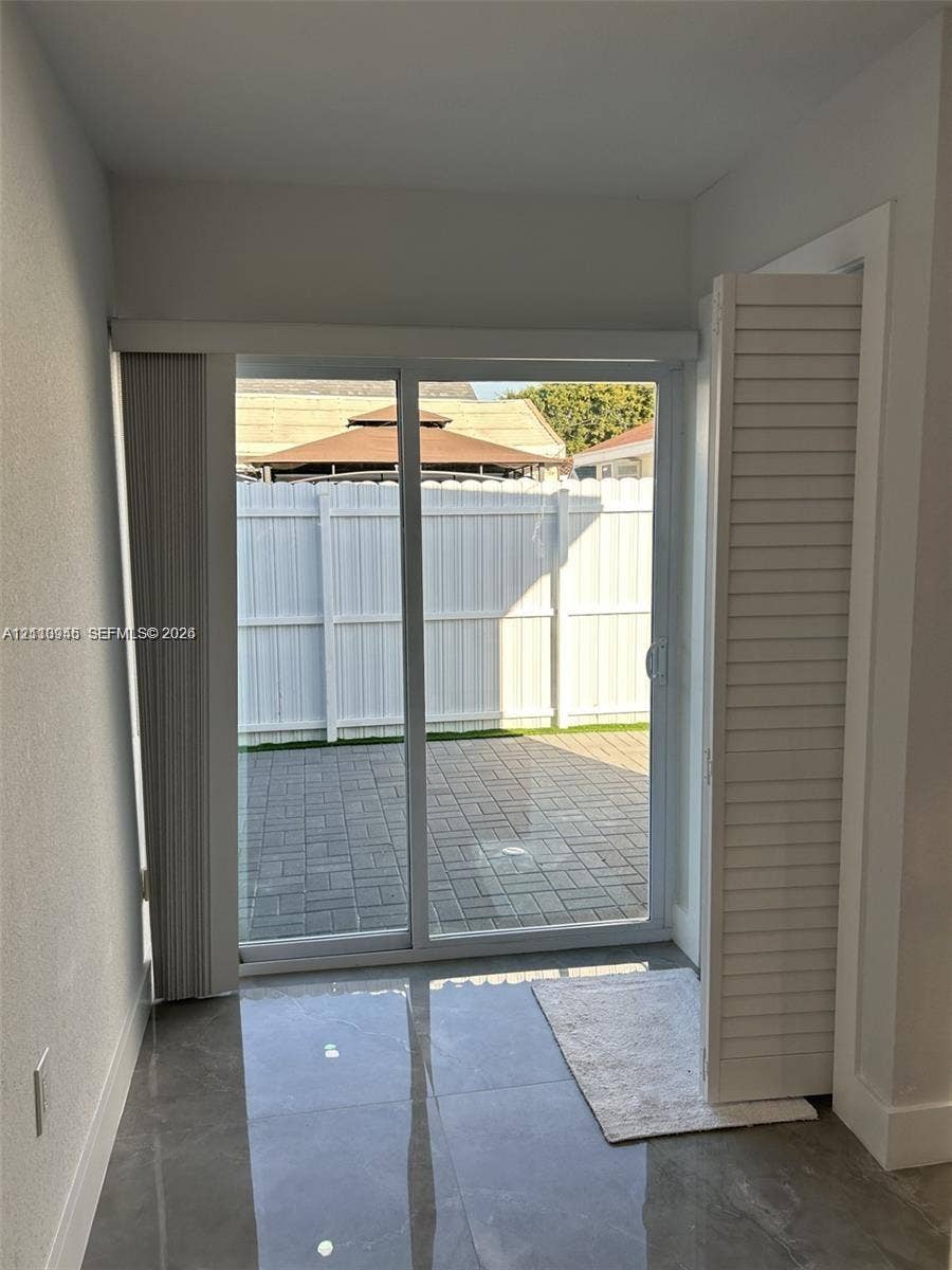 Sliding glass door with vertical blinds, polished large-format tile flooring, and white louvered closet doors opening to a paved patio enclosed by a white privacy fence