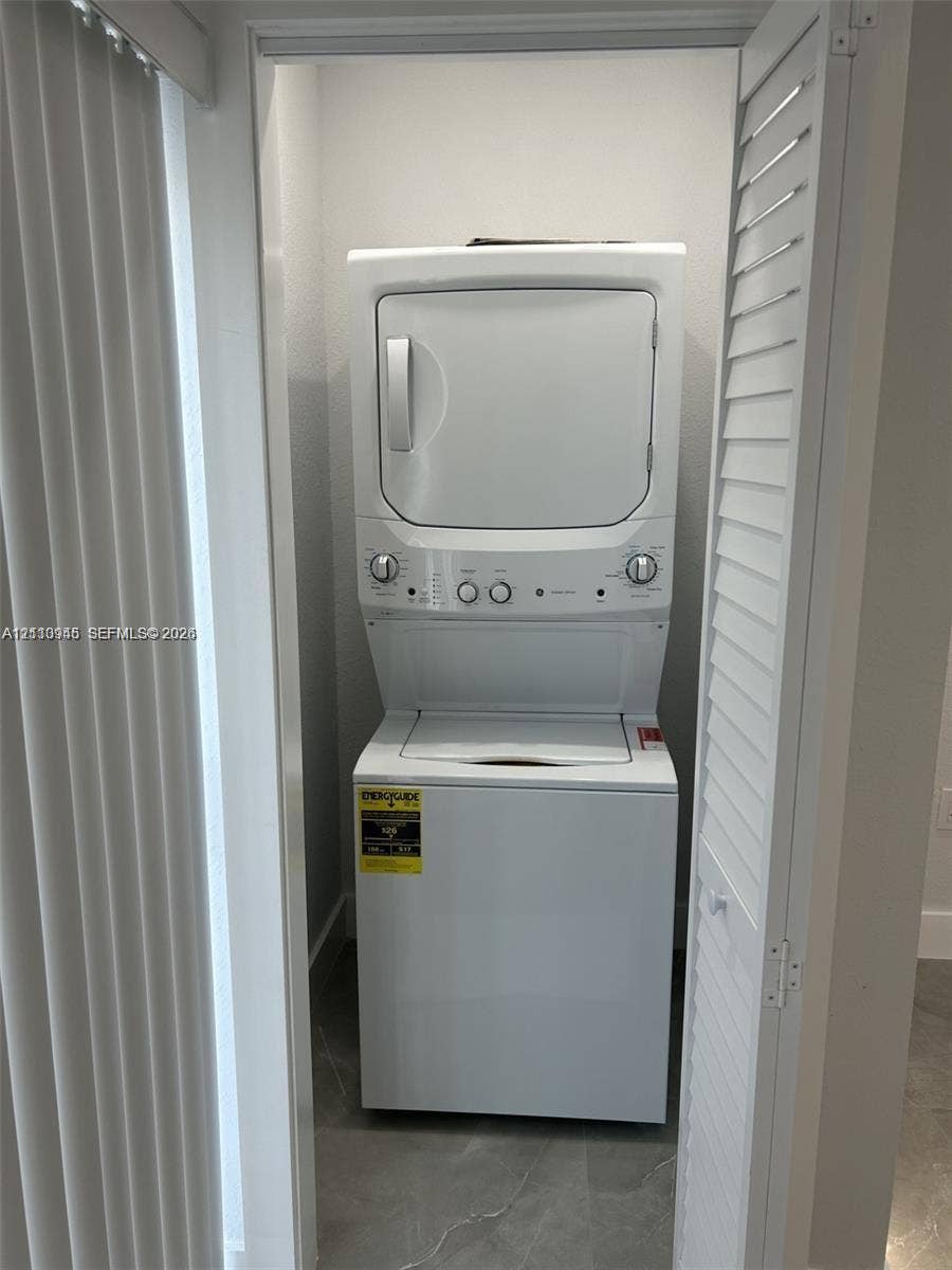 Stacked white laundry unit enclosed by louvered bi-fold doors, featuring tile flooring and vertical blind hardware