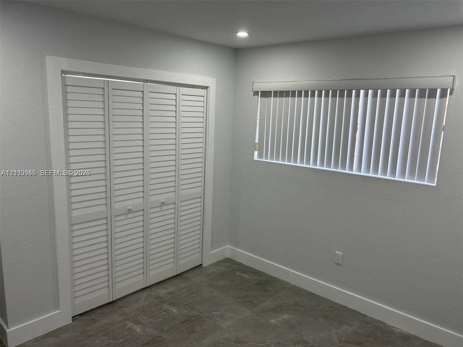 Room featuring neutral-toned walls, tile flooring, louvered bi-fold closet doors, and a window equipped with vertical blinds