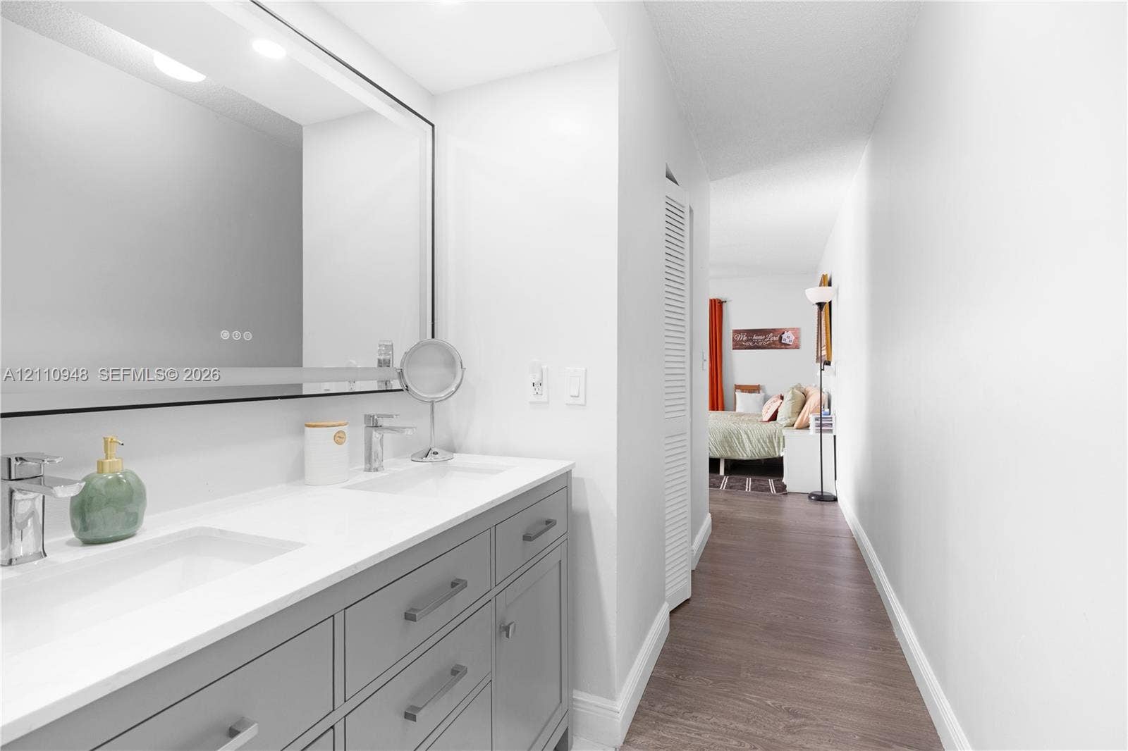 Dual-sink vanity with white stone countertop, integrated rectangular basins, grey cabinetry, and large backlit wall mirror featuring wood-finish flooring