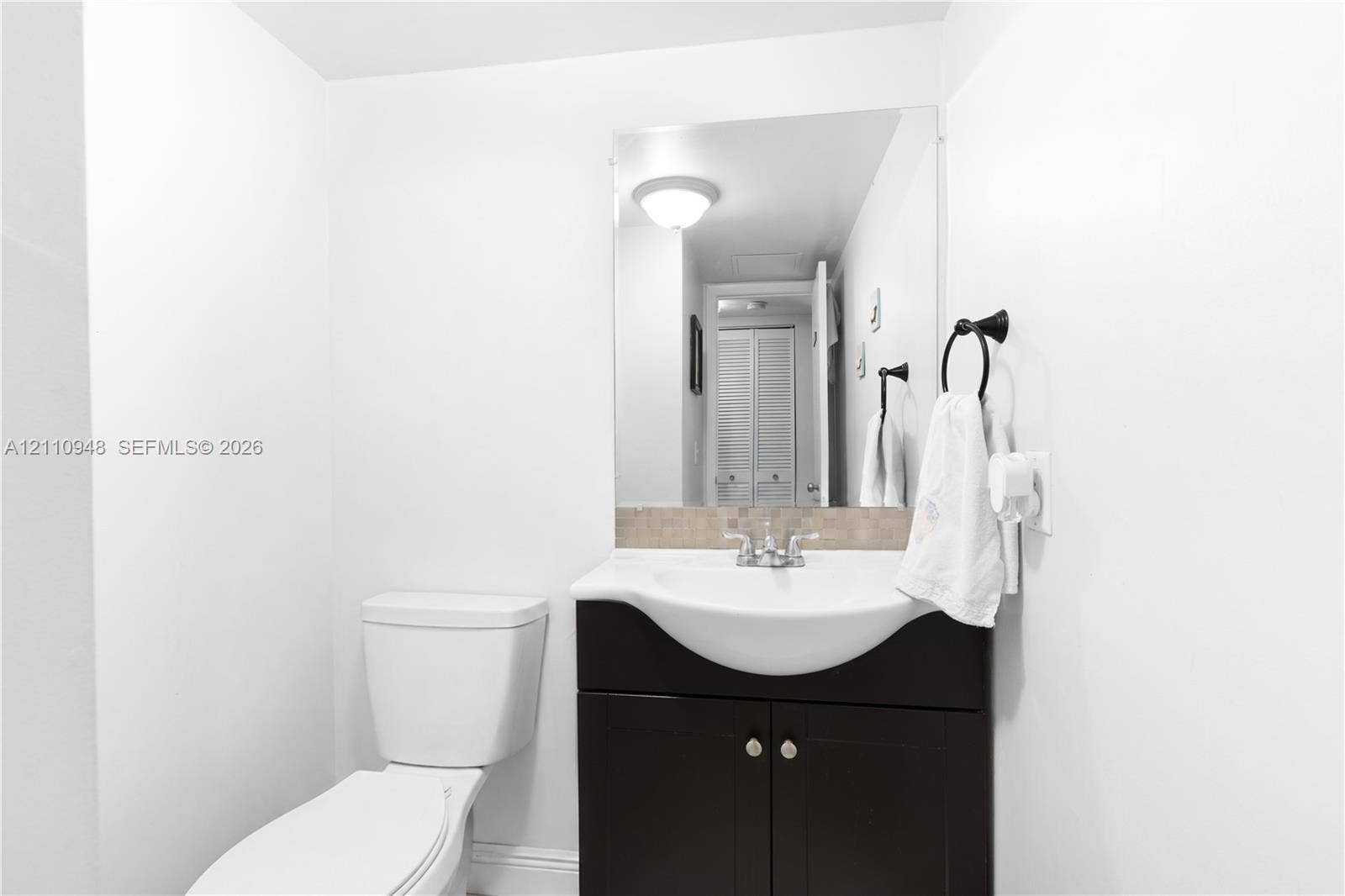 Bathroom vanity featuring a dark-finish base cabinet, integrated white basin, chrome hardware, stone-composite backsplash, and a wall-mounted mirror