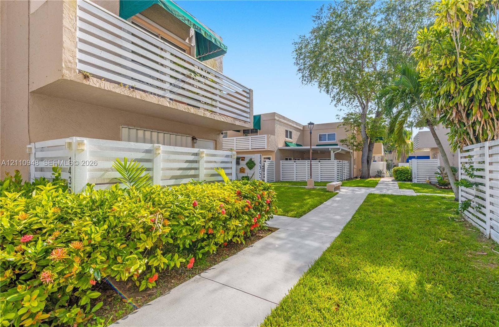 Exterior featuring a concrete walkway, white slatted privacy fencing, second-story balcony with fabric awning, and mature tropical landscaping
