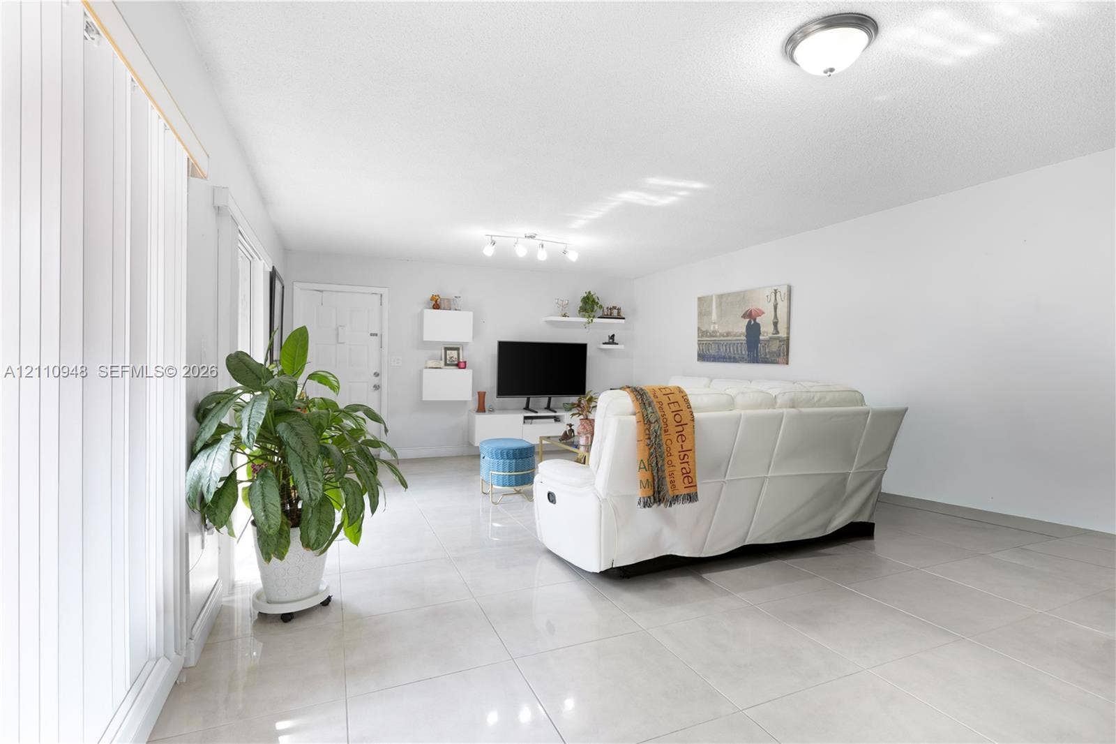 Large-format tile flooring, multi-bulb overhead track lighting, sliding glass door with vertical blinds, and floating wall-mounted shelving