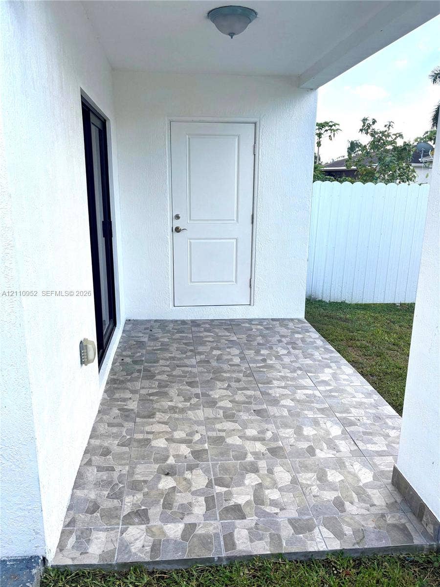1225 NW 9 Avenue, #0, Florida City, Florida City, FL 33034 Covered outdoor patio featuring patterned stone-finish tile flooring, white stucco exterior walls, a white paneled entry door, and a sliding glass door