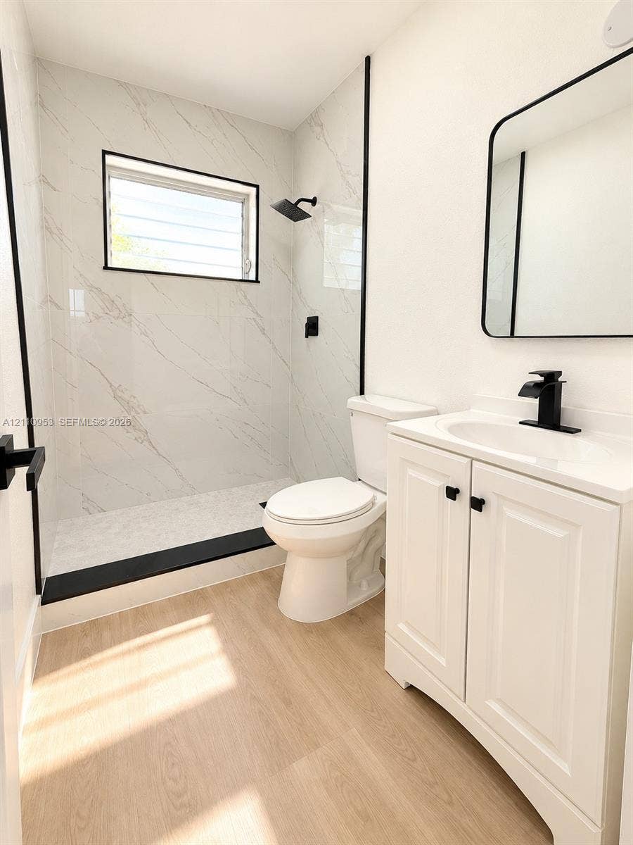 Bathroom featuring walk-in shower with stone-patterned wall surround, wood-finish flooring, white vanity with integrated sink, matte black hardware, and rounded-corner mirror