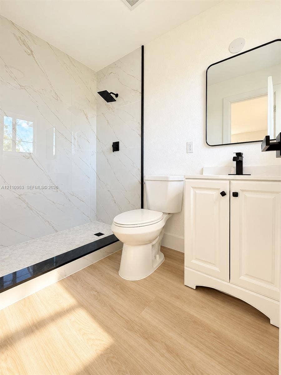 Walk-in shower with stone-finish tile surround, matte black rainfall showerhead, wood-finish flooring, white vanity cabinet with black hardware, and rectangular mirror with black frame