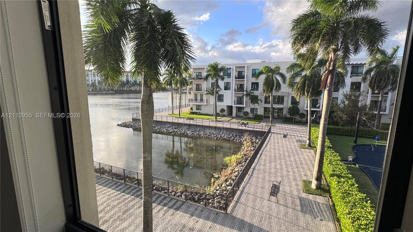 4636 NW 84th Avenue, #35, Doral, FL 33166 Waterfront property featuring a stone-paver walkway, metal perimeter fencing, mature palm trees, and manicured landscaping