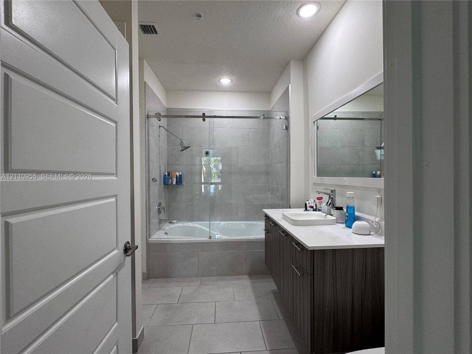 4636 NW 84th Avenue, #35, Doral, FL 33166 Bathroom featuring a vanity with wood-finish cabinetry, white countertop, rectangular sink, full-width mirror, and a tub-shower combination with a sliding glass door and large-format tile surround