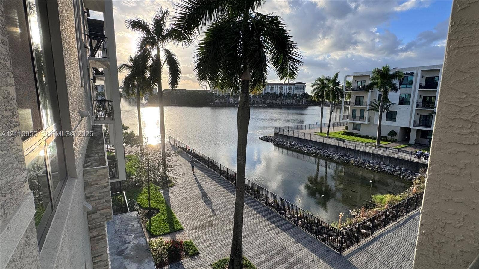 4636 NW 84th Avenue, #35, Doral, FL 33166 Waterfront property featuring a paved stone promenade, metal perimeter fencing, mature palm trees, and structural stucco exterior detailing