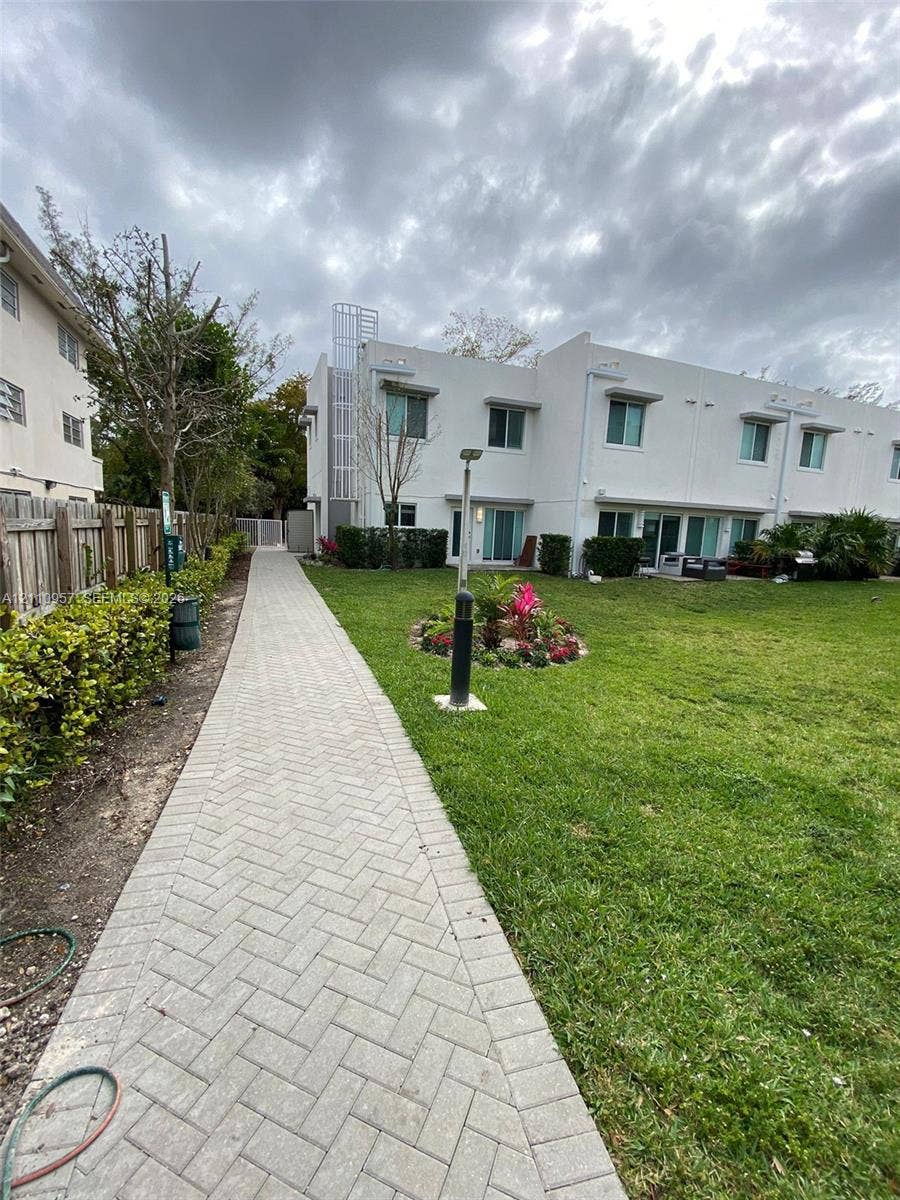 12115 NE 5th Avenue, #12115, North Miami, North Miami, FL 33161 Exterior of a multi-unit property featuring white stucco siding, a herringbone-patterned paver walkway, manicured lawn, established landscaping, and a wood privacy fence