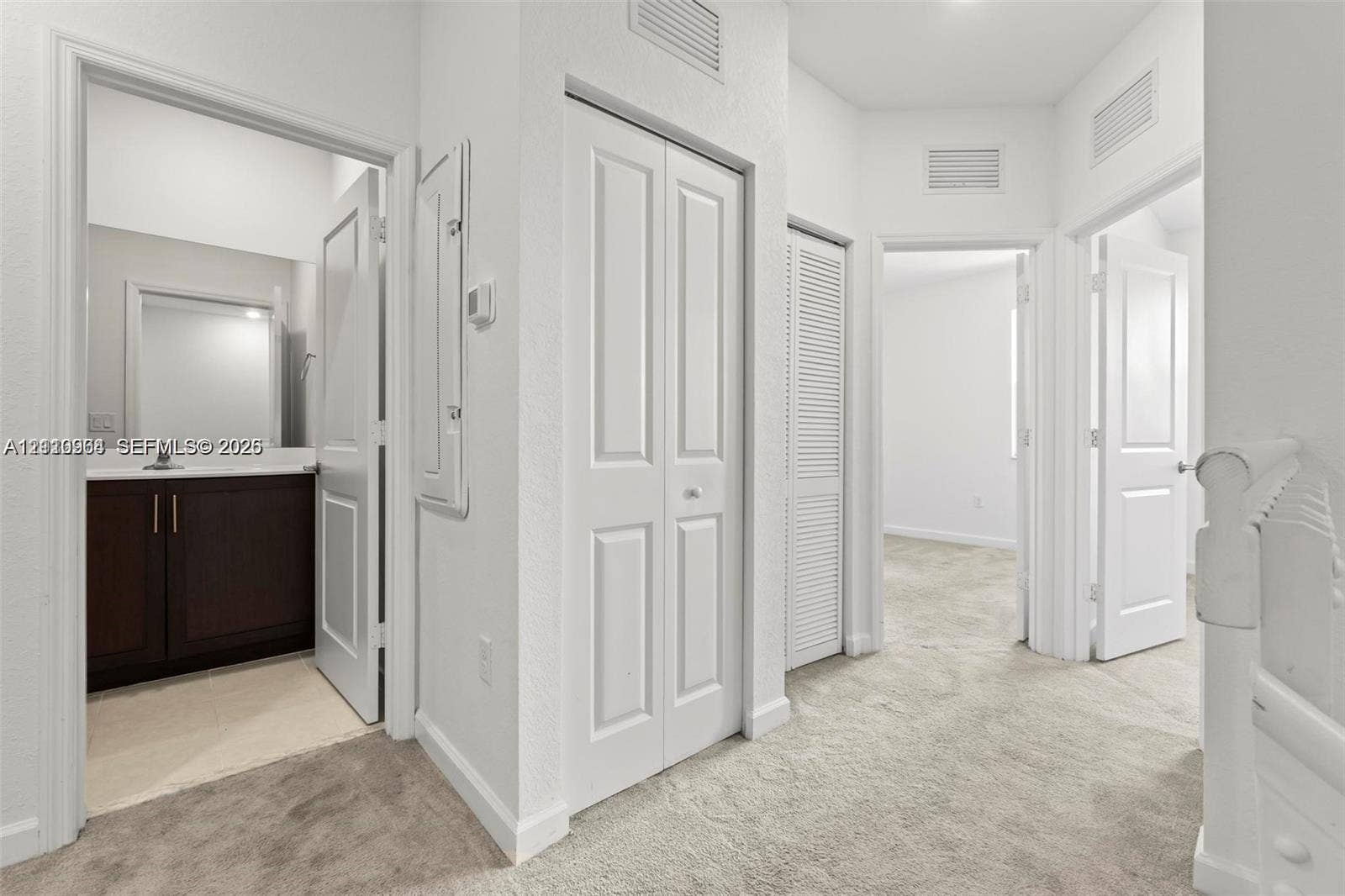 Hallway featuring neutral carpeting, a built-in closet with paneled double doors, a separate louvered door closet, and a bathroom containing a dark-toned vanity cabinet with light-colored countertop