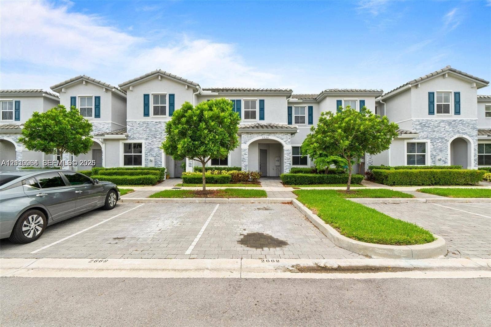 Multi-unit exterior featuring light gray stucco siding with stone veneer accents, arched entryways, and dedicated paver parking spaces