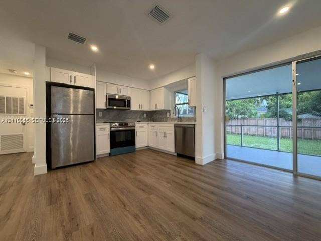 Kitchen featuring white shaker-style cabinetry, stainless steel appliances, stone-finish backsplash, wood-finish flooring, and a large sliding glass door