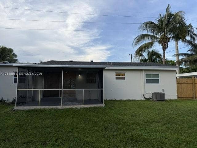 Rear exterior featuring a screened-in lanai, concrete patio, expansive lawn, wood fencing, and mature palm trees
