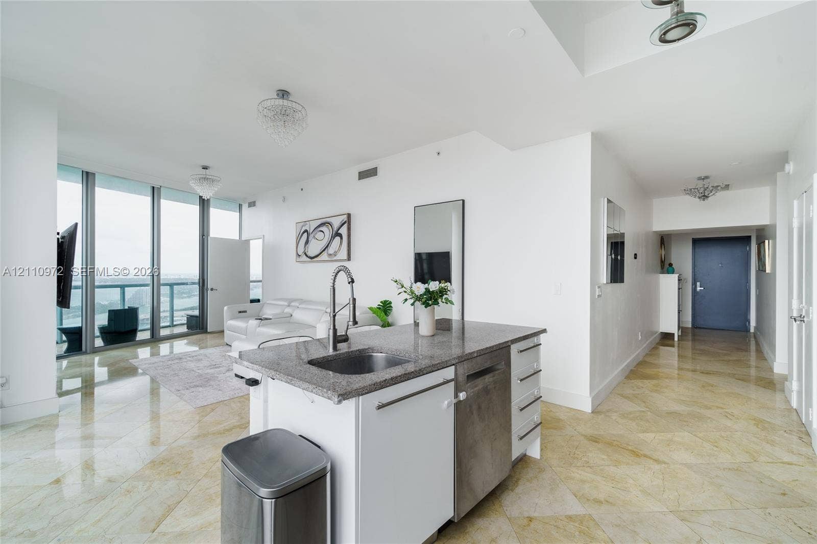 Kitchen island featuring stone-finish countertops, stainless steel undermount sink, integrated dishwasher, and built-in cabinetry, complemented by floor-to-ceiling windows and polished stone-finish flooring