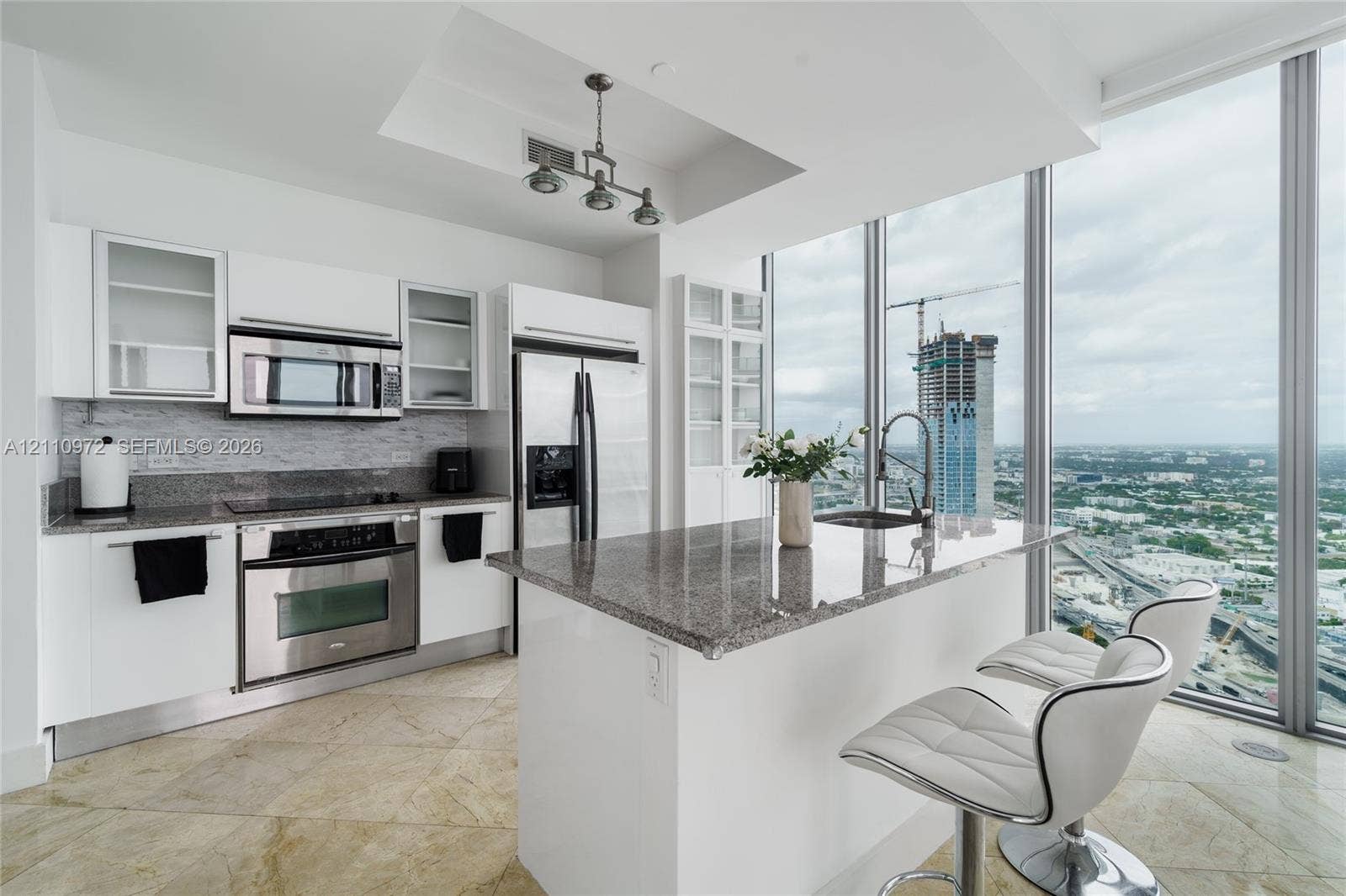 Kitchen featuring custom white cabinetry, stone countertops, stainless steel appliances, a central island with an integrated sink, and floor-to-ceiling windows