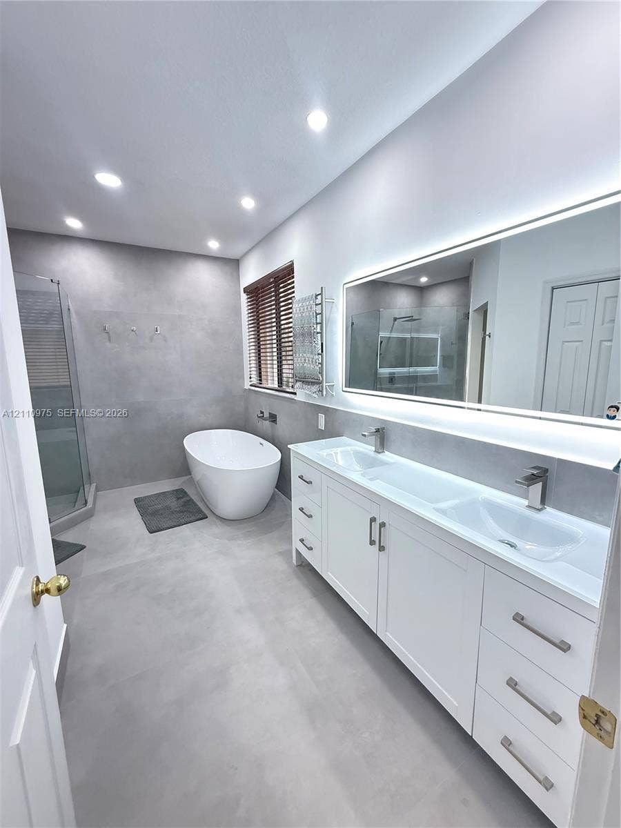 2030 NW 99th Terrace, Pembroke Pines, FL 33024 Dual-vanity cabinet with integrated sinks and metallic hardware, expansive backlit mirror, freestanding soaking tub, enclosed glass shower, and large-format tile flooring